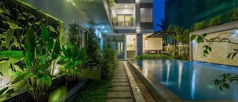A modern building with a swimming pool, surrounded by greenery and a pathway.