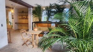 Uma sala de jantar com uma mesa e cadeiras de madeira, rodeada por plantas verdes grandes.