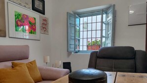 Une pièce avec une table en bois, un fauteuil en cuir noir, un canapé rose agrémenté de coussins couleur moutarde, et une fenêtre donnant sur un paysage verdoyant.