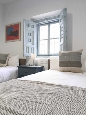 A bedroom with two beds, a nightstand, a lamp, and a window with a view. - Mina25 by 7Escalones (Rota)