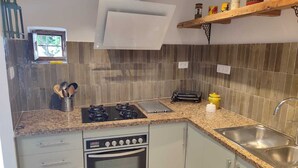 Une cuisine compacte équipée d’une cuisinière à gaz, d’un four et d’un micro-ondes encastré. Une petite fenêtre se trouve au-dessus de l’évier, et une étagère contenant des conserves est placée au-dessus du plan de travail.