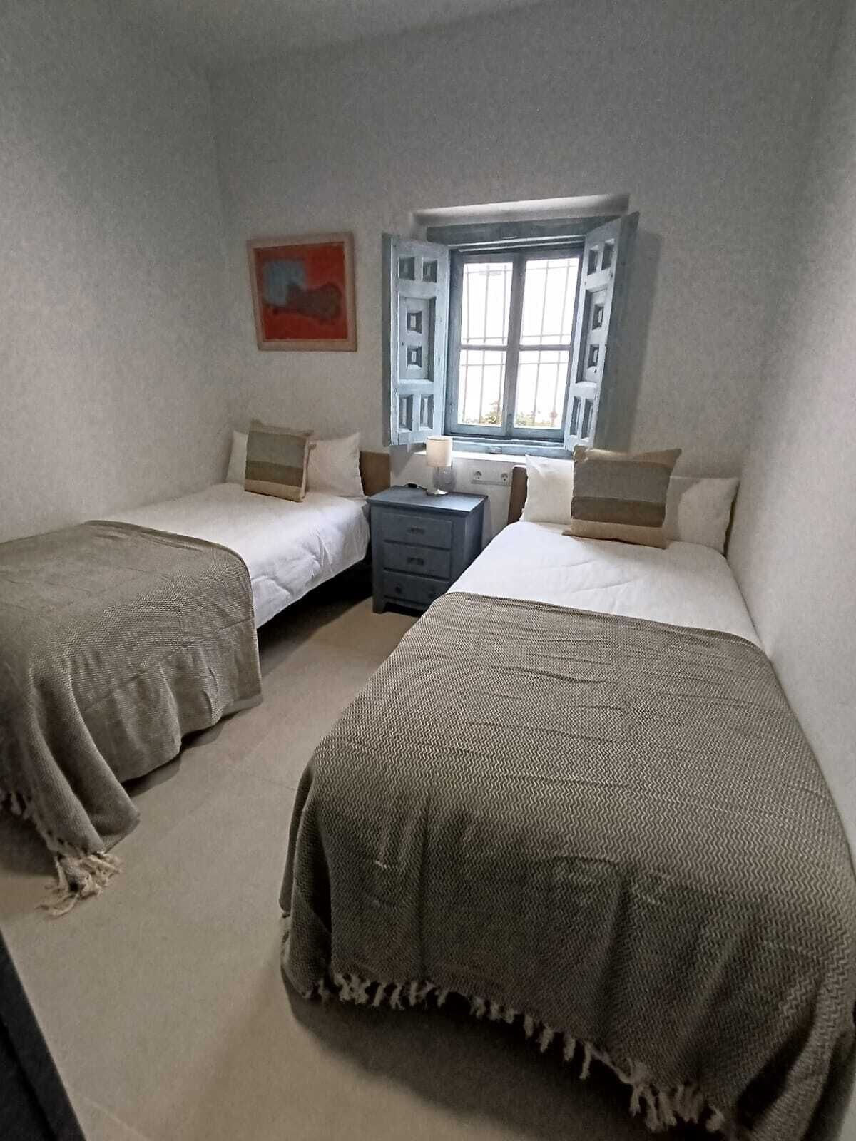 Quarto com duas camas, uma mesinha de cabeceira, janela com persianas e um quadro na parede.