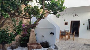 Une cour avec un jardinière en pierre, du gravier blanc, une table et des chaises en bois, et un bâtiment blanc doté d’une porte et d’une fenêtre.