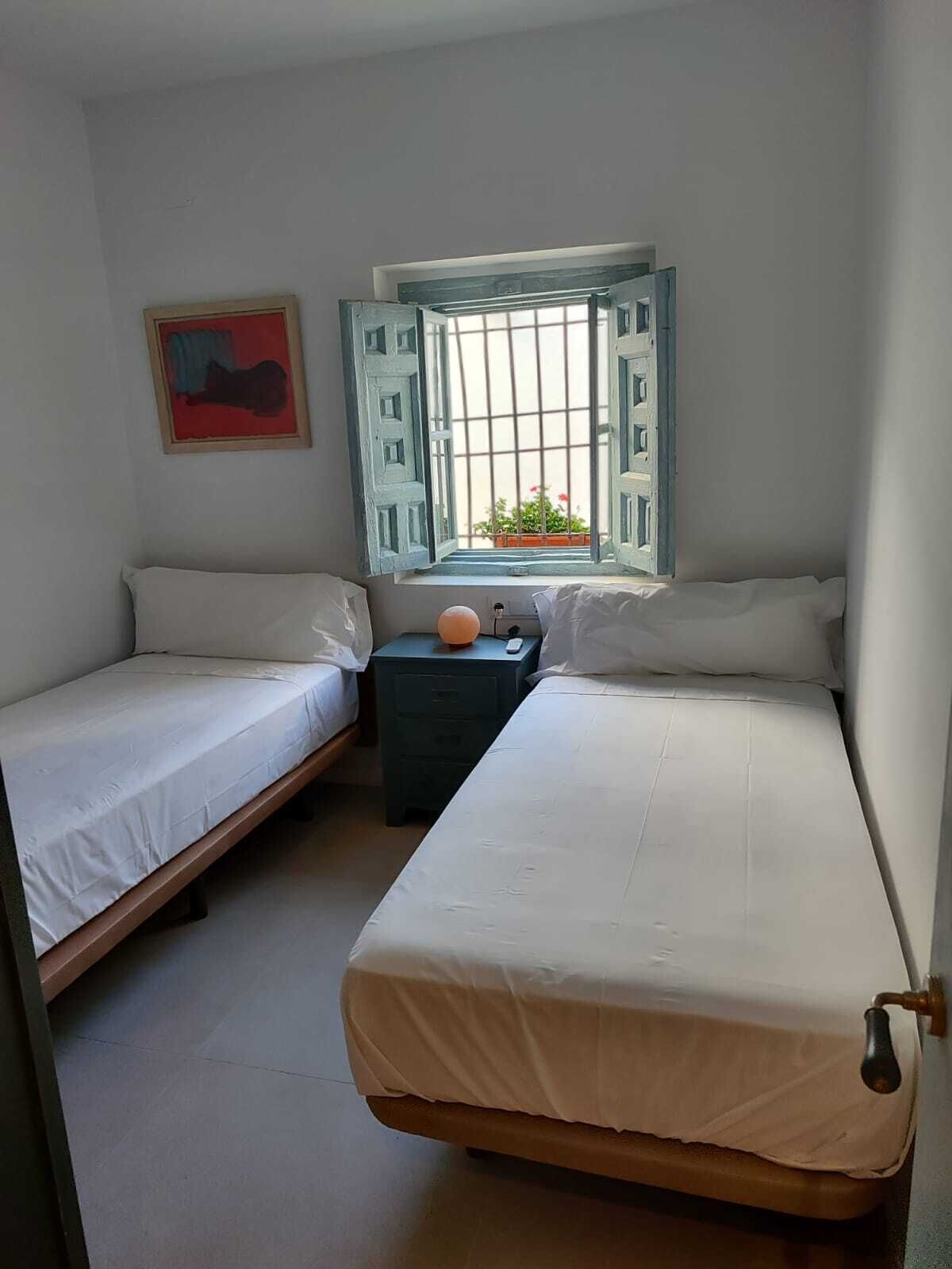 A small, clean room with two single beds, a nightstand, a window with a view of a balcony, and a framed picture on the wall.