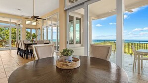 A spacious dining area with a round table, chairs, and a ceiling fan. Large windows offer a view of the ocean.