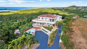 A large, two-story house with a swimming pool, surrounded by lush greenery and a view of the ocean.
