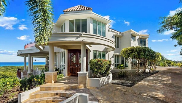 A modern two-story house with a curved balcony, a large wooden door, and a view of the ocean.