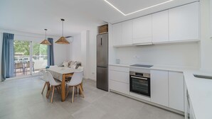 Una cocina moderna con armarios blancos, un refrigerador de acero inoxidable y un horno. Hay una mesa de comedor con sillas y lámparas colgantes.