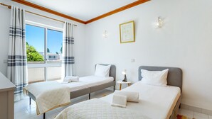 Een hotelkamer met twee bedden, een balkon met gordijnen, een klein tafeltje met een lamp en een lijst met een afbeelding aan de muur.