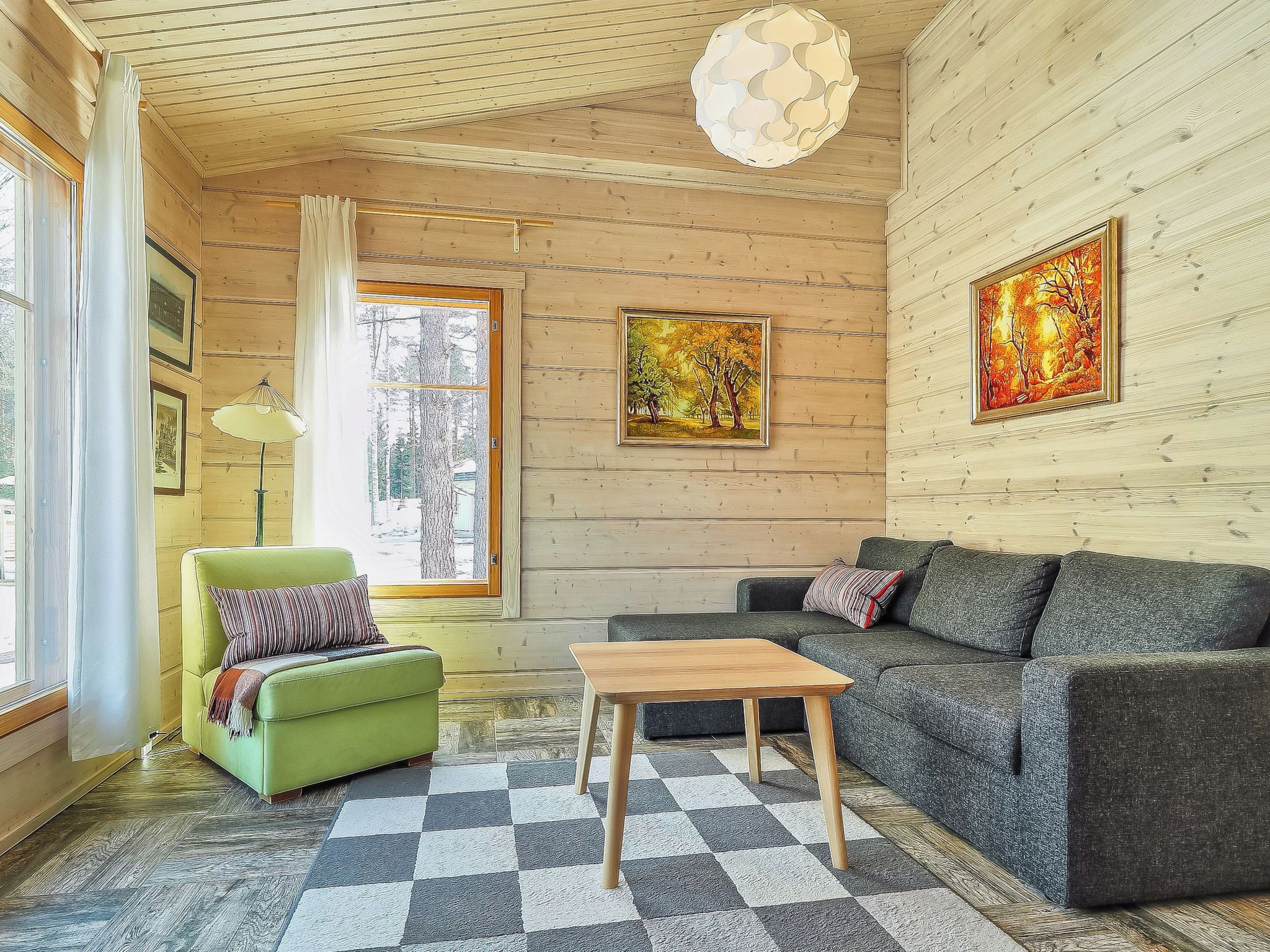 A cozy room with a green armchair, a grey sofa, a wooden coffee table, and a checkered rug. There are framed pictures on the walls and a window with curtains.