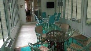 Un patio couvert avec une table et des chaises en verre, un téléviseur fixé au mur et une vue sur l’extérieur.