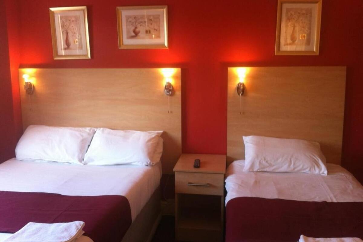 Quarto de hotel com duas camas, cada uma com roupa de cama branca e colchas vermelhas. Há duas mesas de cabeceira com abajures, e as paredes são pintadas de vermelho.