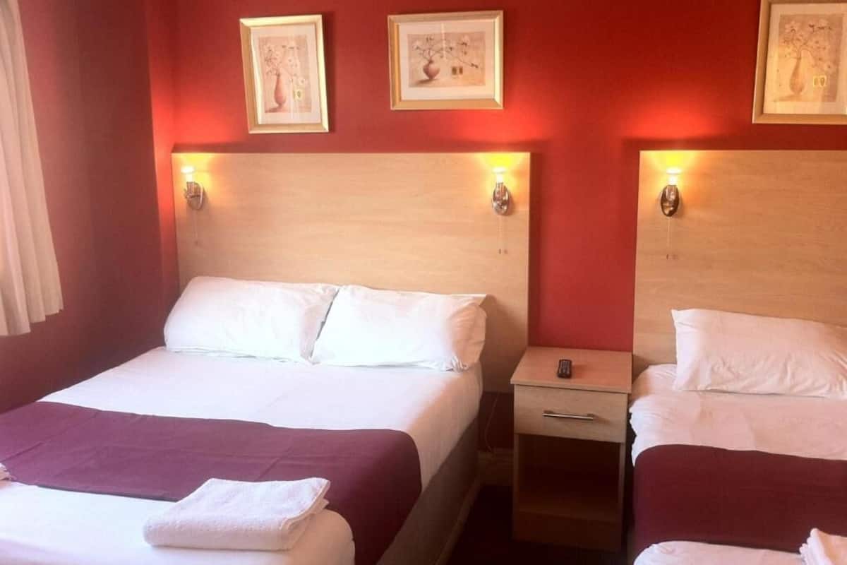 Quarto de hotel com duas camas, cada uma com cabeceira de madeira e luminárias laterais. Quadros emoldurados decoram a parede acima das camas.