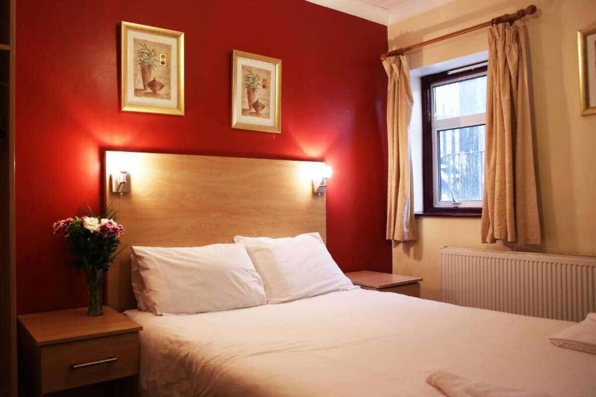 Quarto de hotel com cabeceira de madeira, duas imagens emolduradas na parede, mesa de cabeceira com um vaso de flores e uma janela com cortinas.
