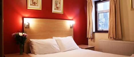 Habitación de hotel con cabecera de madera, dos cuadros en la pared, una mesita de noche con un florero y una ventana con cortinas.