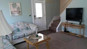A living room with a sofa, a coffee table, a TV on a wooden stand, and a staircase. - Charming 3-bedroom house with AC and WiFi in Ocean Pines (Ocean Pines)