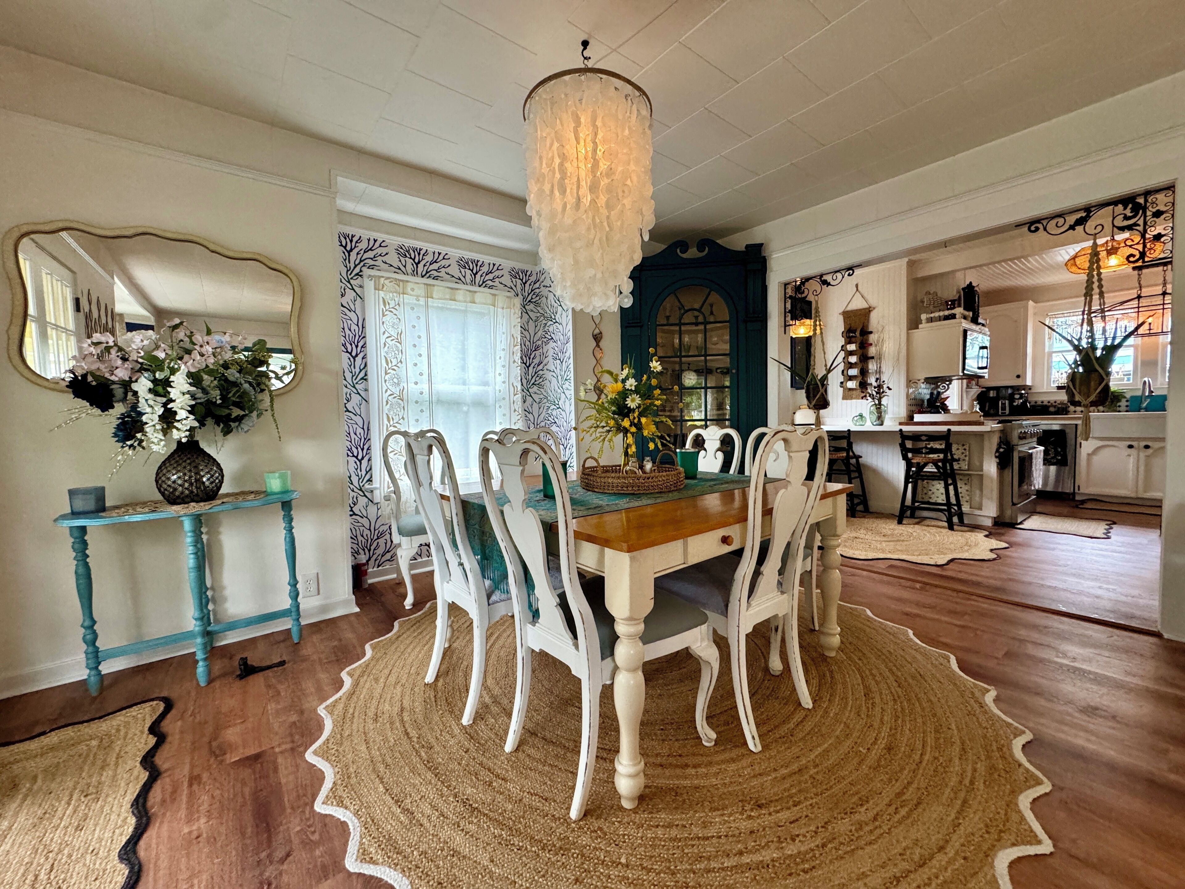 A dining area with a wooden table, white chairs, a chandelier, and a patterned rug.