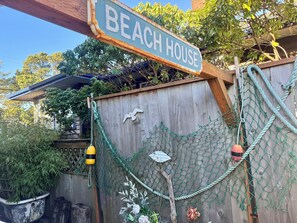 A wooden fence with a 'BEACH HOUSE' sign, a rope net, and decorative elements like a bird and a fish. - Beachy Chic Seaside Retreat-gas fireplace & firepit, steps to beach, prom & town (Seaside)