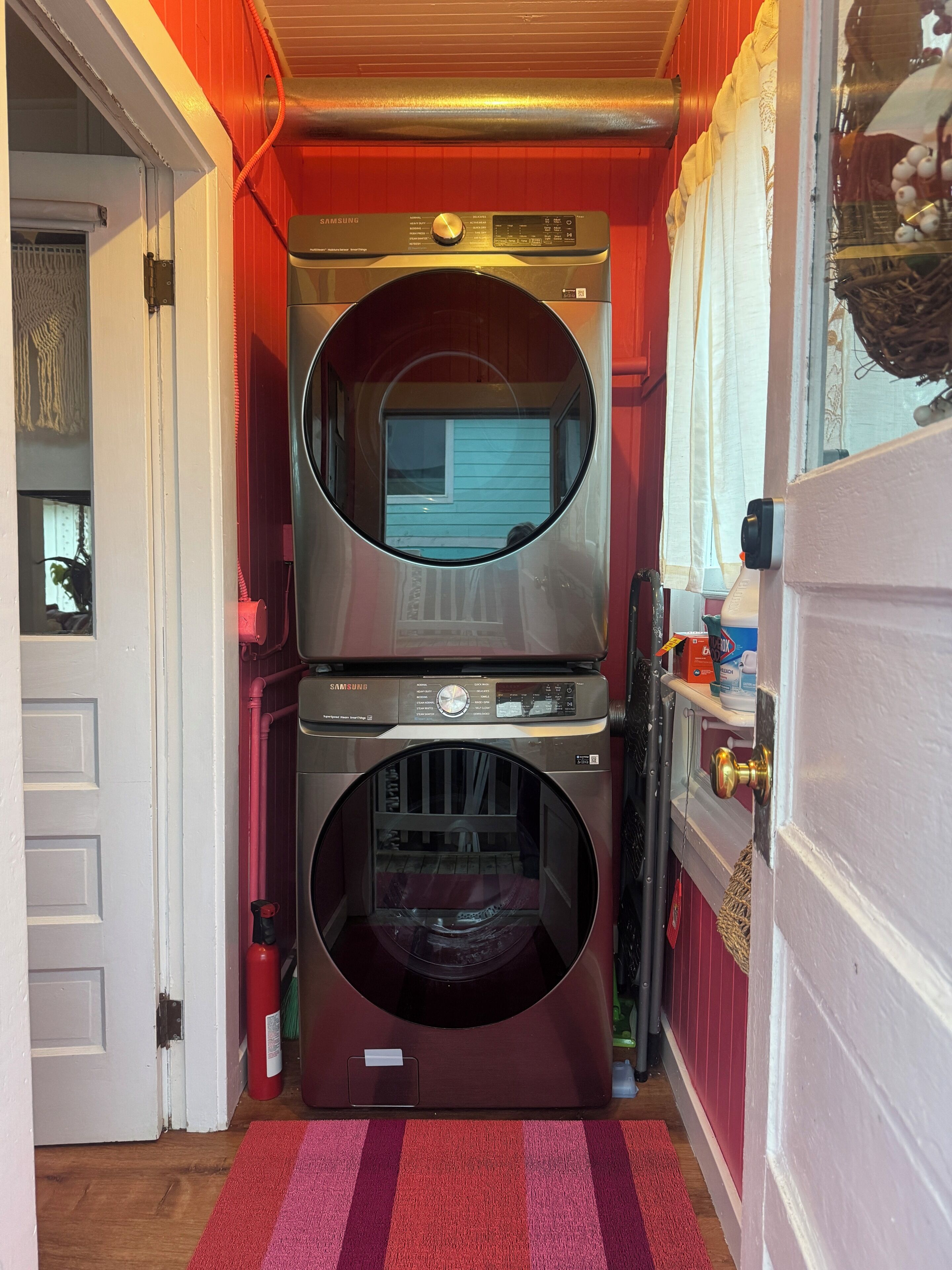 A compact laundry room with a Samsung washer and dryer, a red door, and a small window.