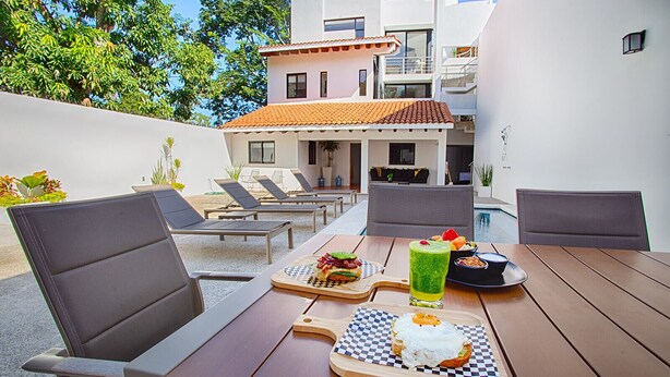 An outdoor dining area with a wooden table, chairs, and a pool in the background. - Brand new modern home w pool- perfect for groups (Sayulita)