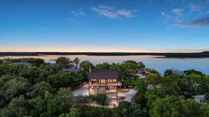 Una casa de dos pisos rodeada de árboles con vista a un lago tranquilo y colinas al lejano.