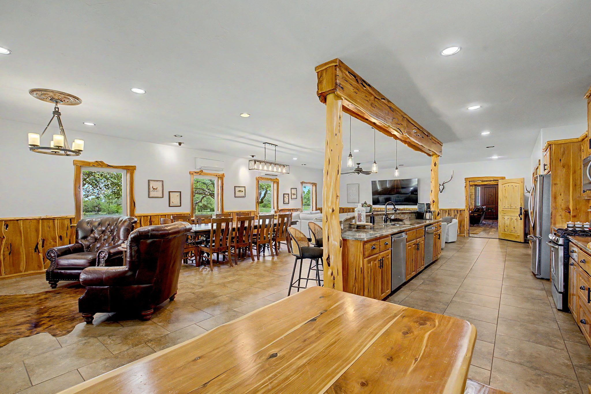 A spacious living area with a large wooden dining table, a leather armchair, and a kitchen with wooden cabinetry and a central island.