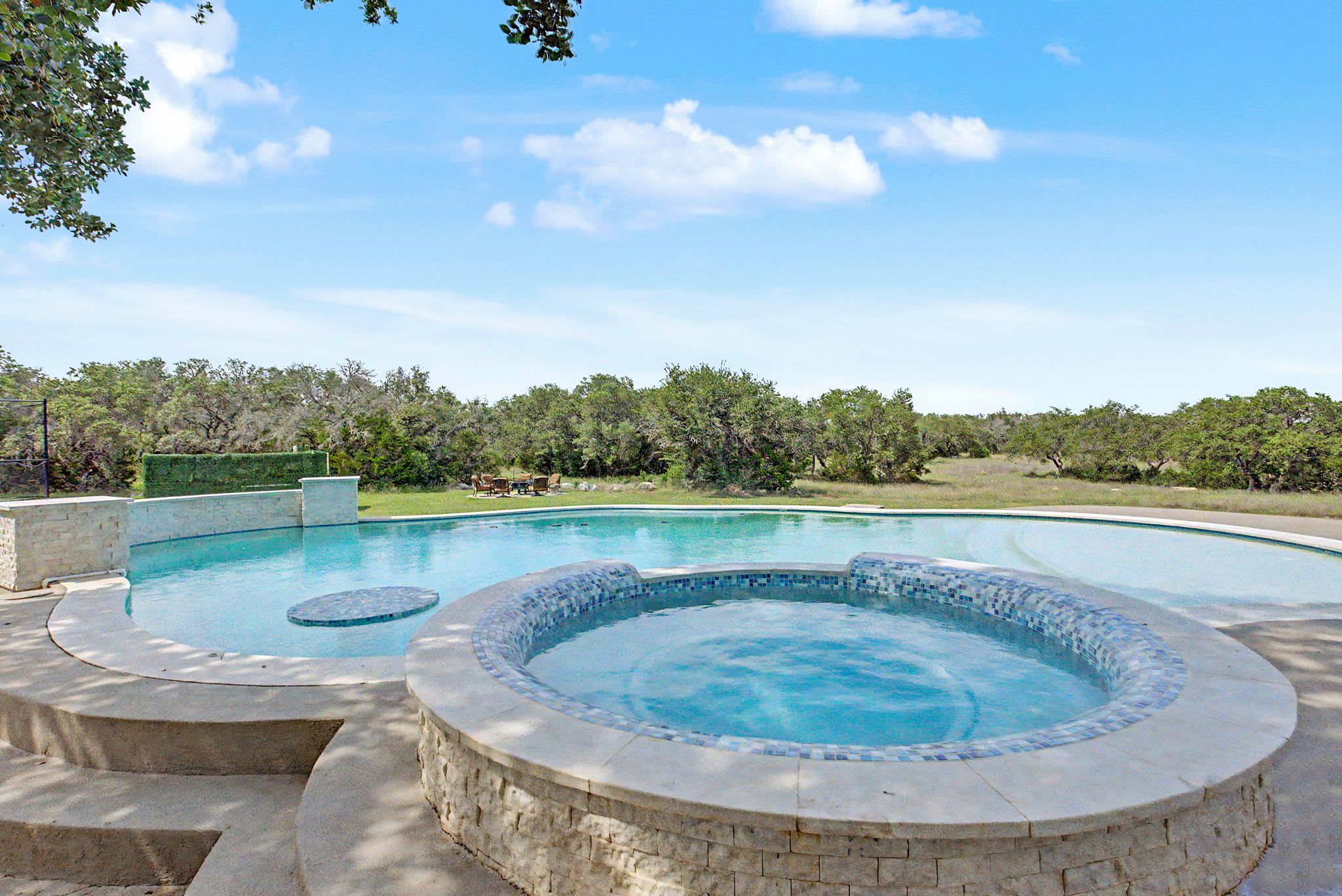 A swimming pool with a hot tub, surrounded by a stone deck and a view of a forested area.