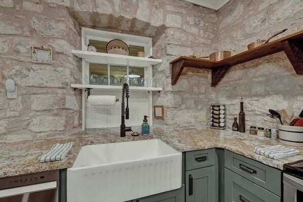 A kitchen with a stone wall, a white sink, and green cabinets. - Historic & Remodeled Sunday Haus-5 Mins to Dtown! (Fredericksburg)