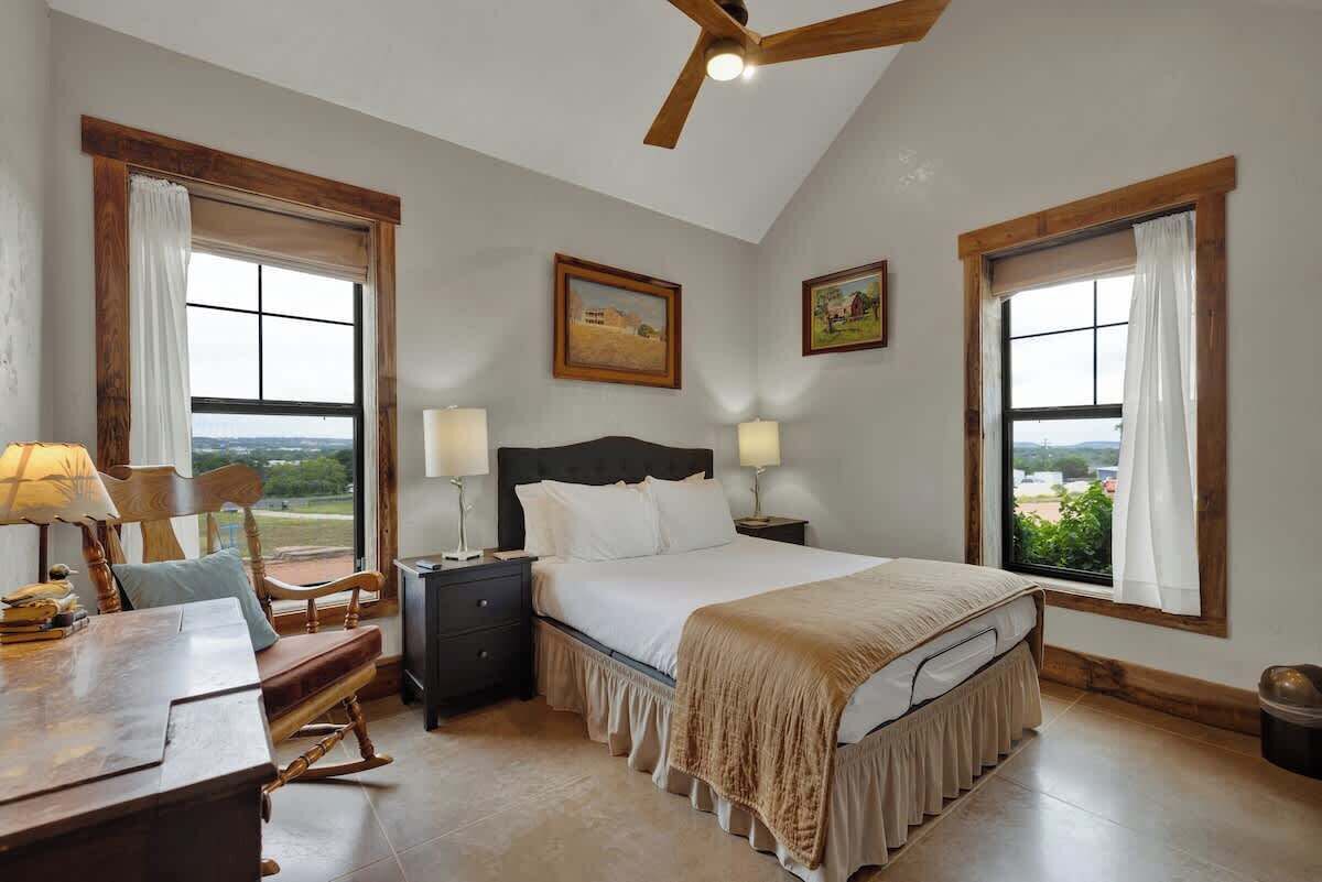 A bedroom with a bed, bedside tables, a desk, and a chair. There are two windows with curtains, a ceiling fan, and a painting on the wall.