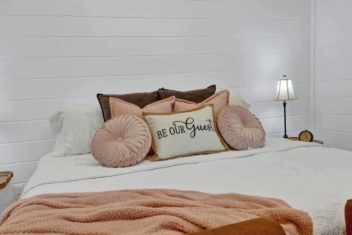 Una cama bien tendida con una almohada que dice 'BE OUR GUEST', una lámpara de noche y un reloj en la mesita.