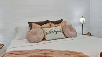 Una cama bien tendida con una almohada que dice 'BE OUR GUEST', una lámpara de noche y un reloj en la mesita.