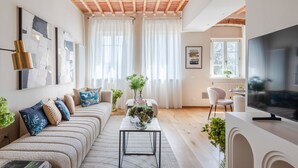 Un salon moderne avec un canapé, une table basse et un téléviseur. On y trouve des rideaux, une plante et des œuvres d’art accrochées aux murs.