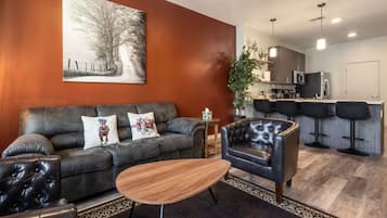 A modern living room with a leather sofa, a wooden coffee table, and a black armchair. A kitchen with bar stools is visible in the background.