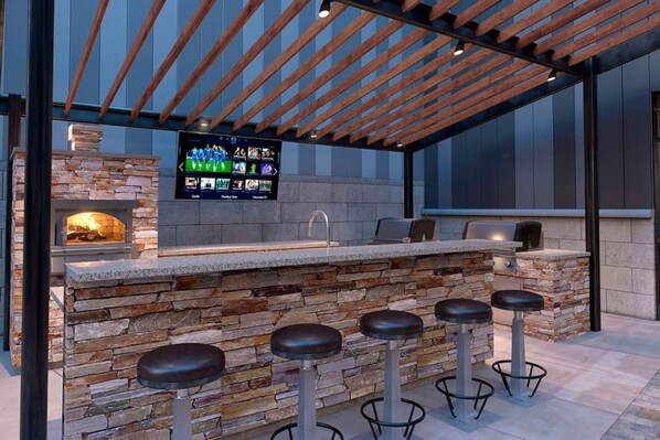 An outdoor bar area with a stone wall, wooden ceiling, and a flat-screen TV mounted above the bar.