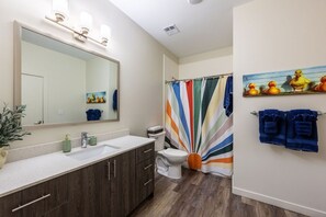 Un baño con un espejo grande, un lavamanos blanco, armarios de madera, un inodoro y una cortina de ducha colorida.