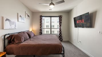 A bedroom with a bed, a ceiling fan, a window with curtains, and a wall-mounted TV displaying the Netflix logo.