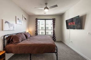 Un dormitorio con una cama, un ventilador de techo, una ventana con cortinas y un televisor de pared que muestra el logotipo de Netflix.