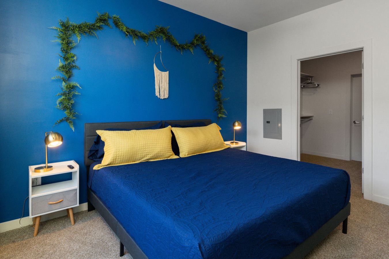 A bedroom with a blue wall, a bed with yellow pillows, a white nightstand with a lamp, and a door leading to another room.