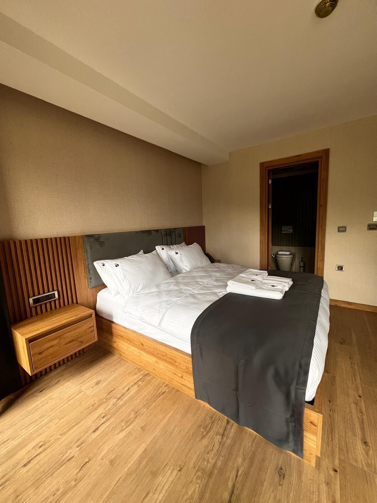 Une chambre d’hôtel moderne avec un plancher en bois, un grand lit avec une literie blanche, une table de chevet et une tête de lit en bois.