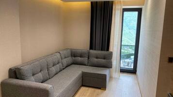 Una sala de estar moderna con un sofá seccional gris, un ventanal con vista a follaje, y una puerta corrediza que da a un espacio exterior.