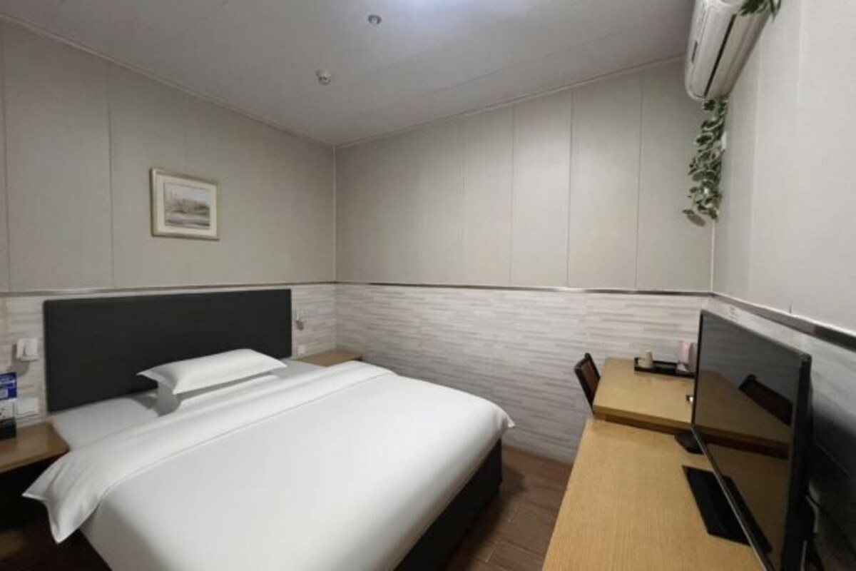 A hotel room with a bed, a desk, a chair, a TV, and an air conditioner.