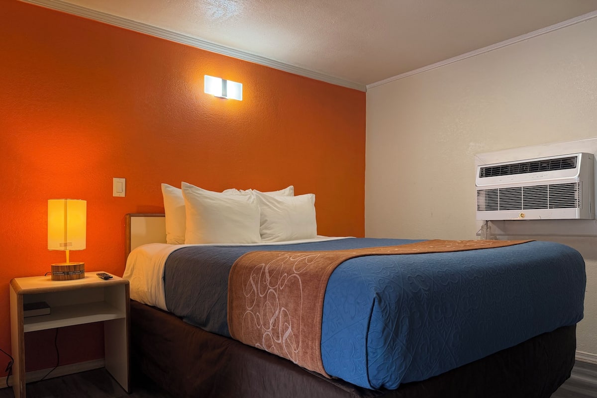 A hotel room with a bed, a nightstand, a lamp, a remote control, and an air conditioning unit.