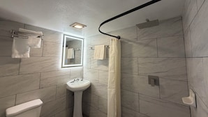 A bathroom with a pedestal sink, a toilet, a shower with a curtain, and a mirror with a light. - Ponca Motel (Abilene)