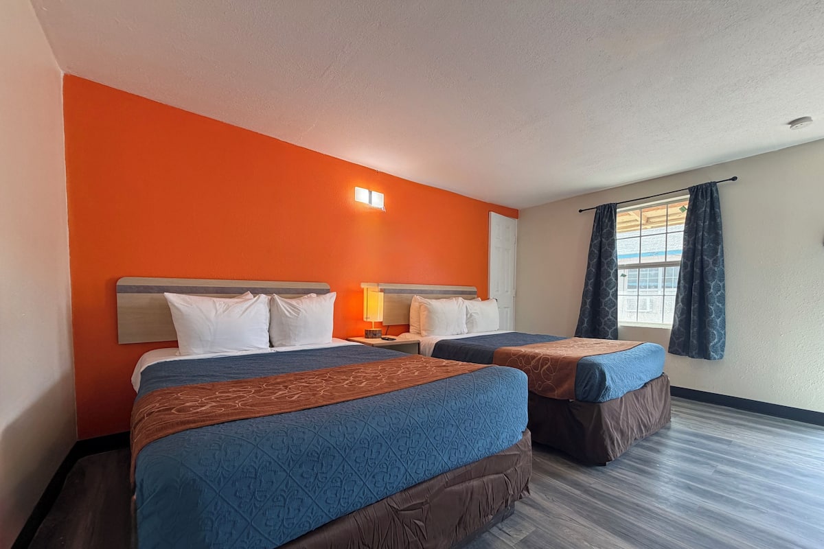 A hotel room with two beds, a nightstand with a lamp, a window with curtains, and a wooden floor.