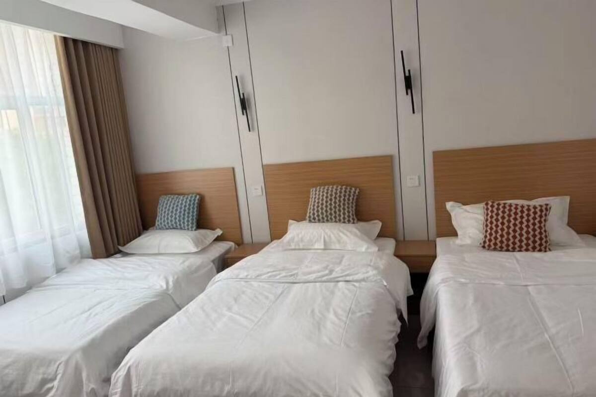 Une chambre d’hôtel avec trois lits simples, une tête de lit en bois et une grande fenêtre avec des rideaux.