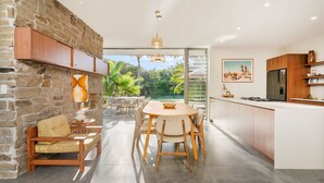 A modern kitchen with a dining area, stone wall, and a view of greenery. - Hikari House - Kingscliff Beachfront (Kingscliff)