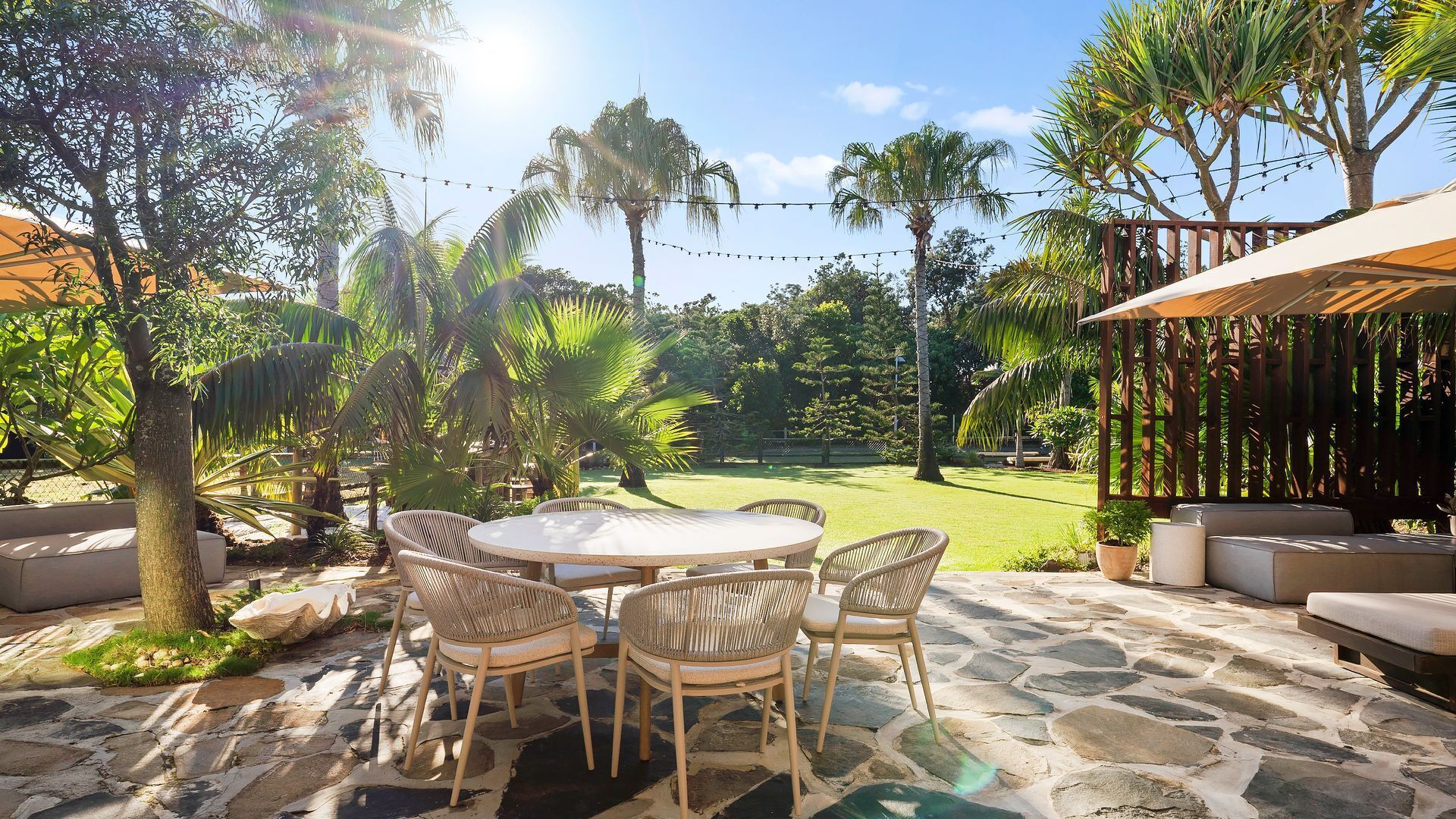 An outdoor patio with a round table and wicker chairs, surrounded by stone flooring and palm trees.