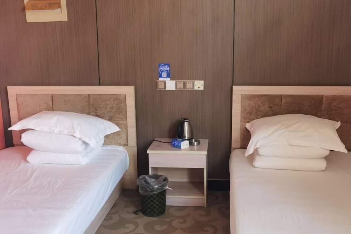 Two adjacent hotel beds with white linens, a small bedside table with a kettle and a trash bin, and a wall-mounted electrical outlet.