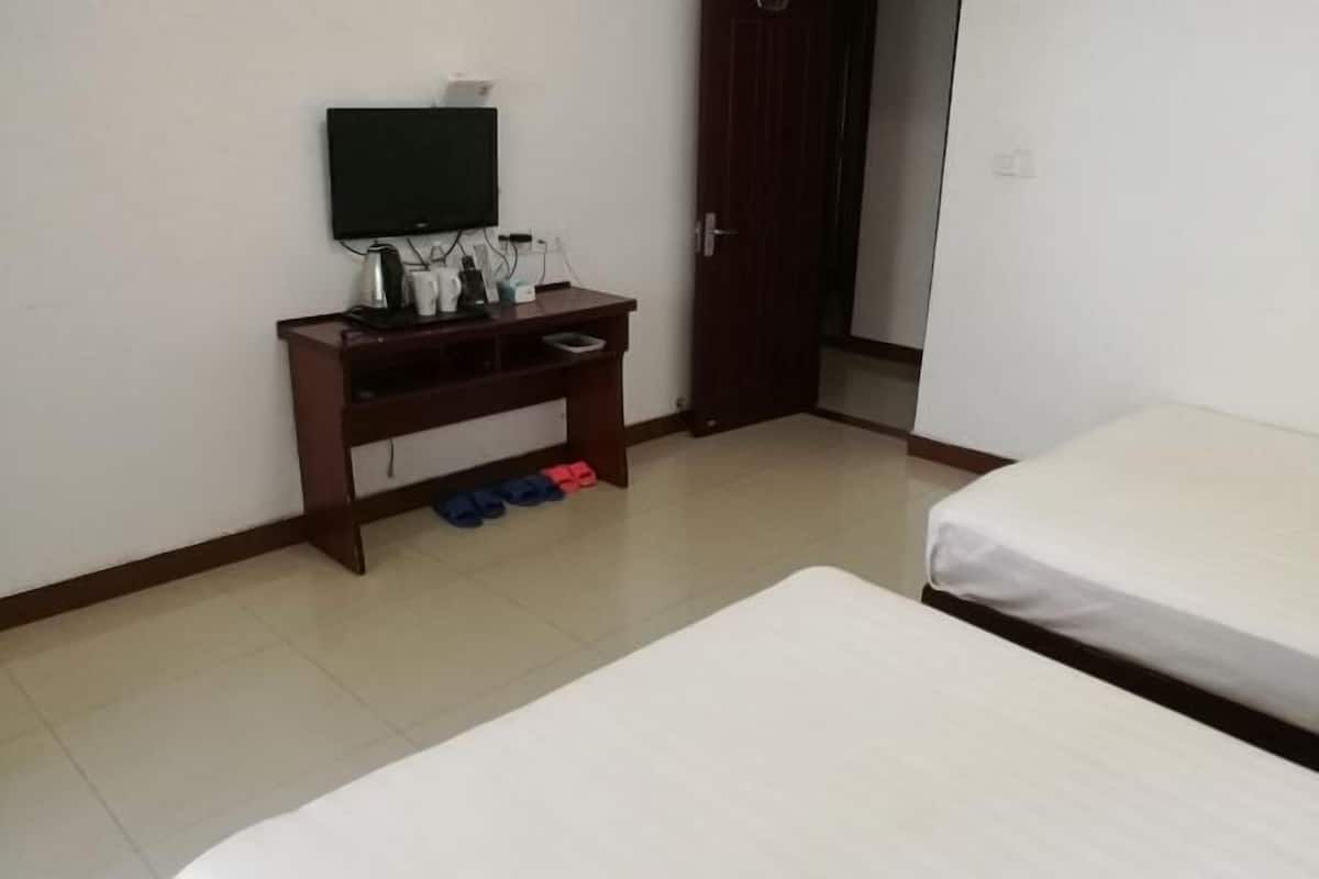 A room with a bed, a TV on a stand, a wooden desk with a kettle, and a door.