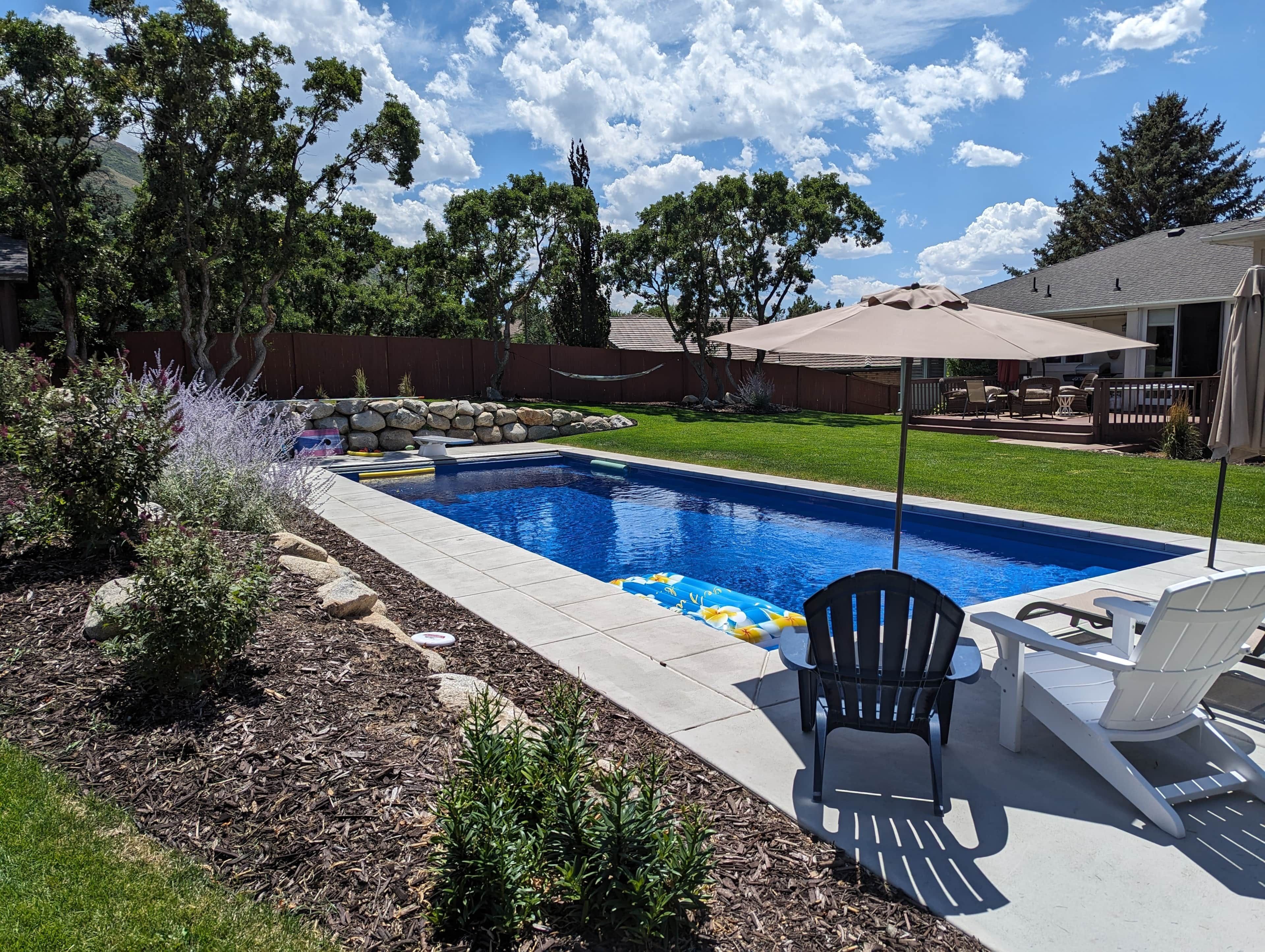 A backyard with a swimming pool, a patio with chairs, and a stone wall.